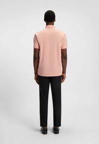 Ljusrosa pikétröja med korta ärmar, platt krage och smal passform, kombinerad med svarta byxor, sedd från baksidan. Slät tygtextur.