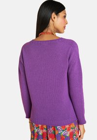 Frau, die sich von hinten zeigt, trägt einen lila Strickpullover, einen Blumenrock, eine rote Perlenkette und große goldene Creolen mit bunten Perlen.