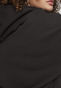 Felpa nera con una texture morbida, caratterizzata da una vestibilità rilassata e dettagli di cucitura visibili sulle cuciture. Orecchini a cerchio dorati sono parzialmente visibili.