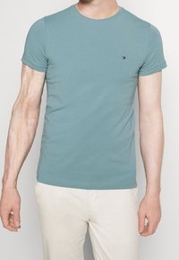 Světle modré bavlněné tričko s krátkým rukávem, kulatým výstřihem a malým vyšitým logem na levé části hrudi. Hladká textura, přiléhavý design.