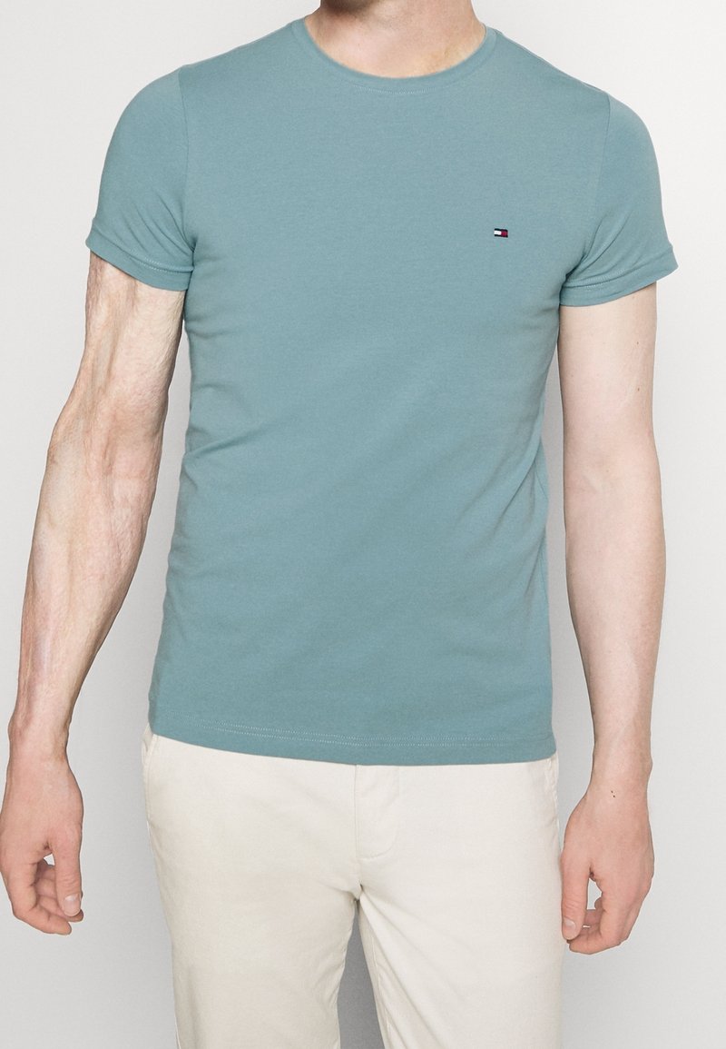 Světle modré bavlněné tričko s krátkým rukávem, kulatým výstřihem a malým vyšitým logem na levé části hrudi. Hladká textura, přiléhavý design.