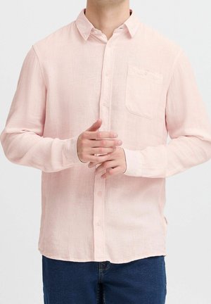 Mand iført lyserød langærmet skjorte med knapper og brystlomme samt mørkeblå jeans, hænderne foldet foran.