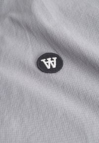 Grijze stof met een rasterstructuur, met een zwarte ronde patch waarop witte gestileerde "AW" letters zijn afgebeeld.