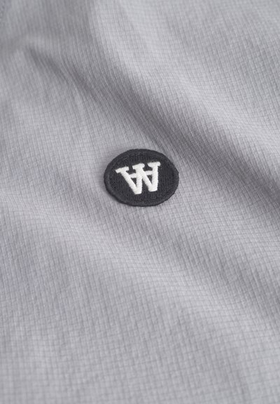 Tissu gris avec une texture en grille présentant un patch circulaire noir avec un lettrage stylisé "AW" en blanc.