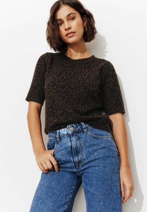 Femme aux cheveux courts et foncés portant un pull à manches courtes à imprimé léopard foncé et un jean bleu taille haute sur fond blanc.
