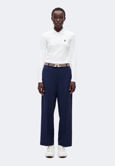 Femme debout, face à nous, portant une chemise blanche à manches longues avec col, un pantalon large bleu marine, une ceinture beige et des baskets blanches sur un fond clair.