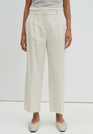 Person trägt hochtaillierte, weit geschnittene, cremefarbene Hosen und passende cremefarbene spitze Ballerinas, steht vor einem schlichten weißen Hintergrund.