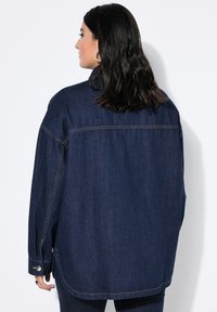 Donkerblauwe denimjack met een relaxte pasvorm, zichtbare stiksels en een platte kraag. Heeft knopen bij de manchetten en zijsplitten in de zoom.