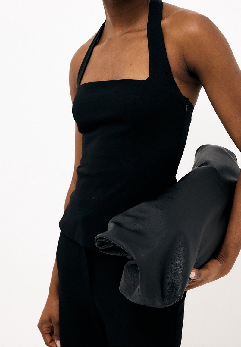 Personne portant un haut dos nu noir tenant une grande pochette en cuir noir roulée sous le bras sur un fond blanc uni.