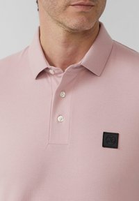 Polo shirt rose pâle avec un tissu texturé, trois boutons blancs et un écusson logo carré noir sur la zone de la poitrine gauche.