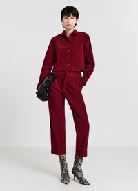 Camicia corta in velluto a coste color bordeaux con bottoni, pantaloni a vita alta abbinati e stivali con tacco a stiletto stampa serpente, pochette nera in mano.