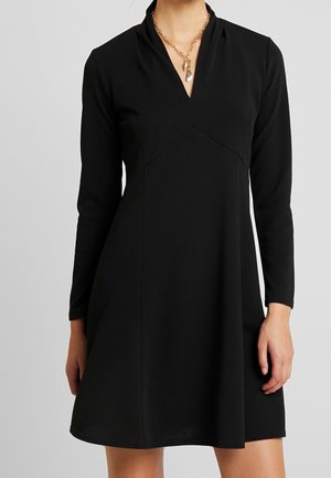 Kobieta w czarnej sukience z długim rękawem i dekoltem w kształcie litery V, nosząca złoty łańcuszek z dwoma wisiorkami na szarym tle.