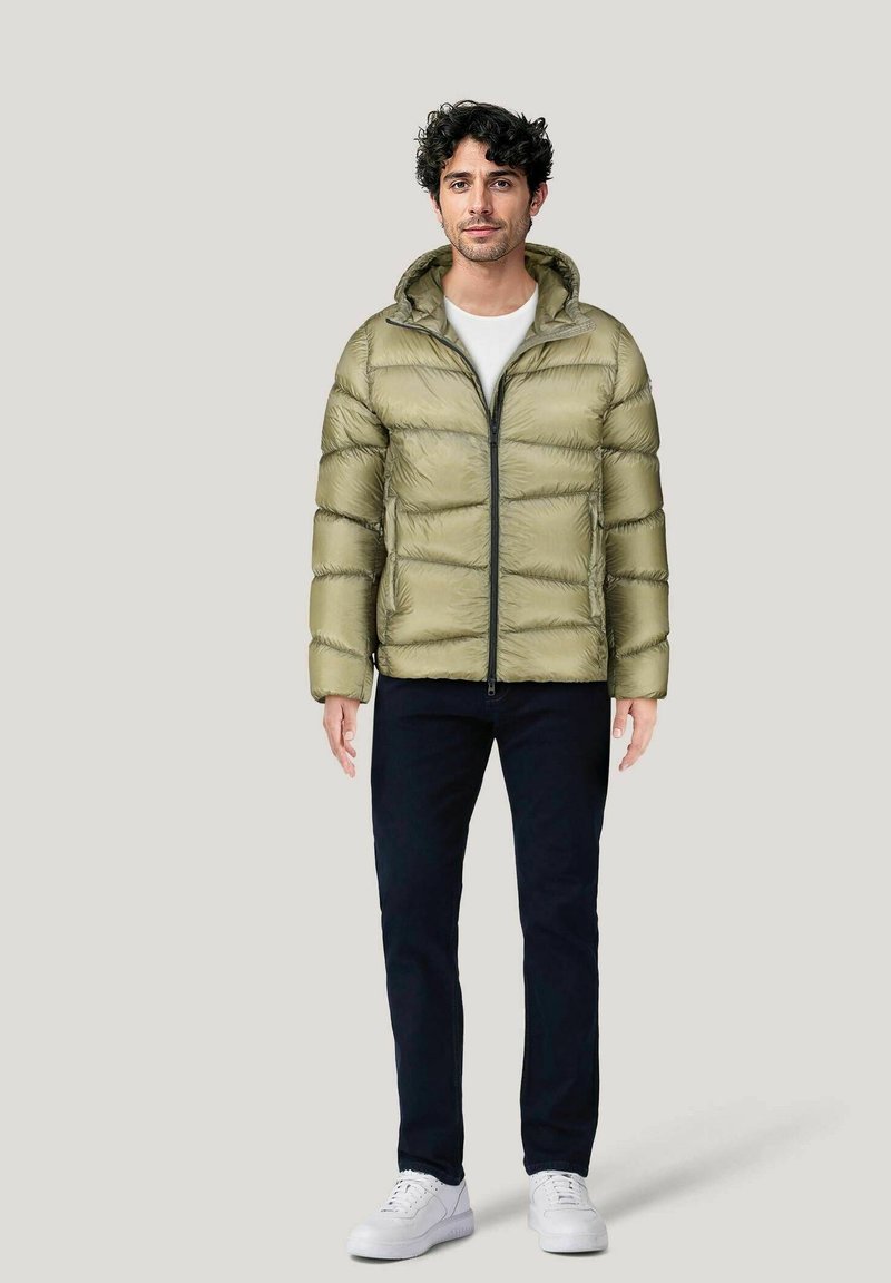 Giacca imbottita verde oliva con zip frontale, cappuccio e finitura texture, abbinata a pantaloni scuri e sneaker bianche. Design minimalista.
