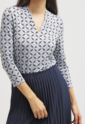 Langærmet bluse i lyseblå med sort geometrisk mønster og V-hals; sat sammen med en navy plisseret nederdel. Blødt stof, tætsiddende design.