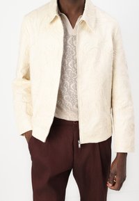 Krämfärgad broderad jacka med en strukturerad krage och dragkedjeslutning, bärs över en texturerad stickad topp och matchas med mörkbruna byxor.