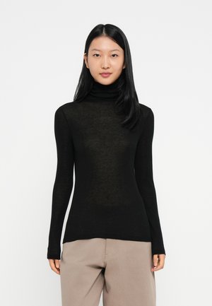 Sieviete, tērpusies melnā garā piedurknē turtleneck džemperī un bēšās biksēs, stāv pretī vienkāršai baltai fona.