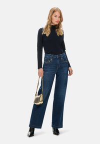 Weite blaue Jeans aus Denim mit detaillierten, mit Nieten versehenen Taschen, kombiniert mit einem figurbetonten schwarzen Langarmoberteil und einer kleinen metallic-goldenen Handtasche.