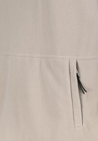 Beiger Fleece-Stoff mit horizontaler Naht und vertikaler Reißverschlusstasche mit schwarzen Zugbändern auf der rechten Seite.