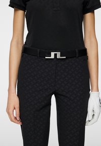 Svart polotröja, mönstrade svarta byxor, svart bälte med silverfärgad geometrisk spänne, och vit golfhandske på vänster hand.