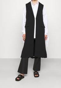 Zwarte lange vest met ingesneden revers en voorzakken, gedragen over een wit overhemd met knopen, gecombineerd met wijde zwarte broekspijpen en zwarte sandalen.