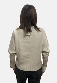 Persona con capelli castani lunghi fino alle spalle, indossa un maglione beige chiaro con polsini e orlo a coste, abbinato a jeans neri, di spalle.
