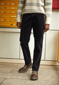 Mörka kordbyxor med rak passform, tillsammans med bruna snörkängor med metallöglor och gummisålar. Randig tröja synlig.