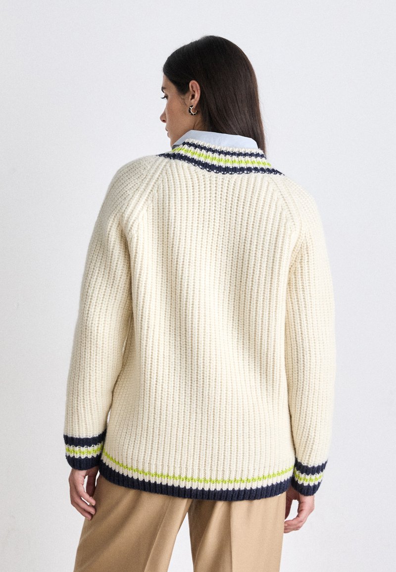 Pull en tricot crème avec une texture côtelée, des rayures contrastées bleu marine et vert citron au niveau du col et des poignets, avec une coupe décontractée.