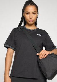 Ung kvinde med fletninger bærer en sort Puma t-shirt med mønstrede kanter og har en sort crossbody-taske på skulderen.