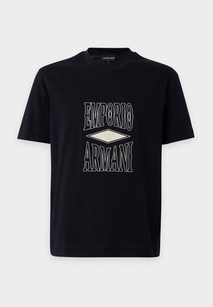 Νέος άνδρας που φοράει μαύρο T-shirt Emporio Armani, μαύρο παντελόνι και άσπρα αθλητικά παπούτσια, στέκεται με το ένα χέρι στην τσέπη σε λευκό φόντο.
