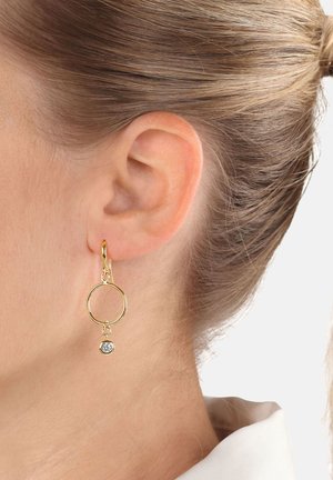 Nahaufnahme eines Ohres, das einen goldenen Creolenohrring mit einem kleinen diamantähnlichen Edelstein, der innerhalb der Creole hängt, trägt. Die Haare sind nach hinten gebunden, ein weißer Kragen ist sichtbar.