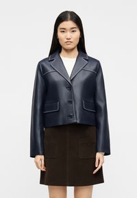 Femme portant une veste en cuir bleu marine boutonnée avec des coutures blanches sur un haut blanc et une jupe marron foncé avec des poches avant.