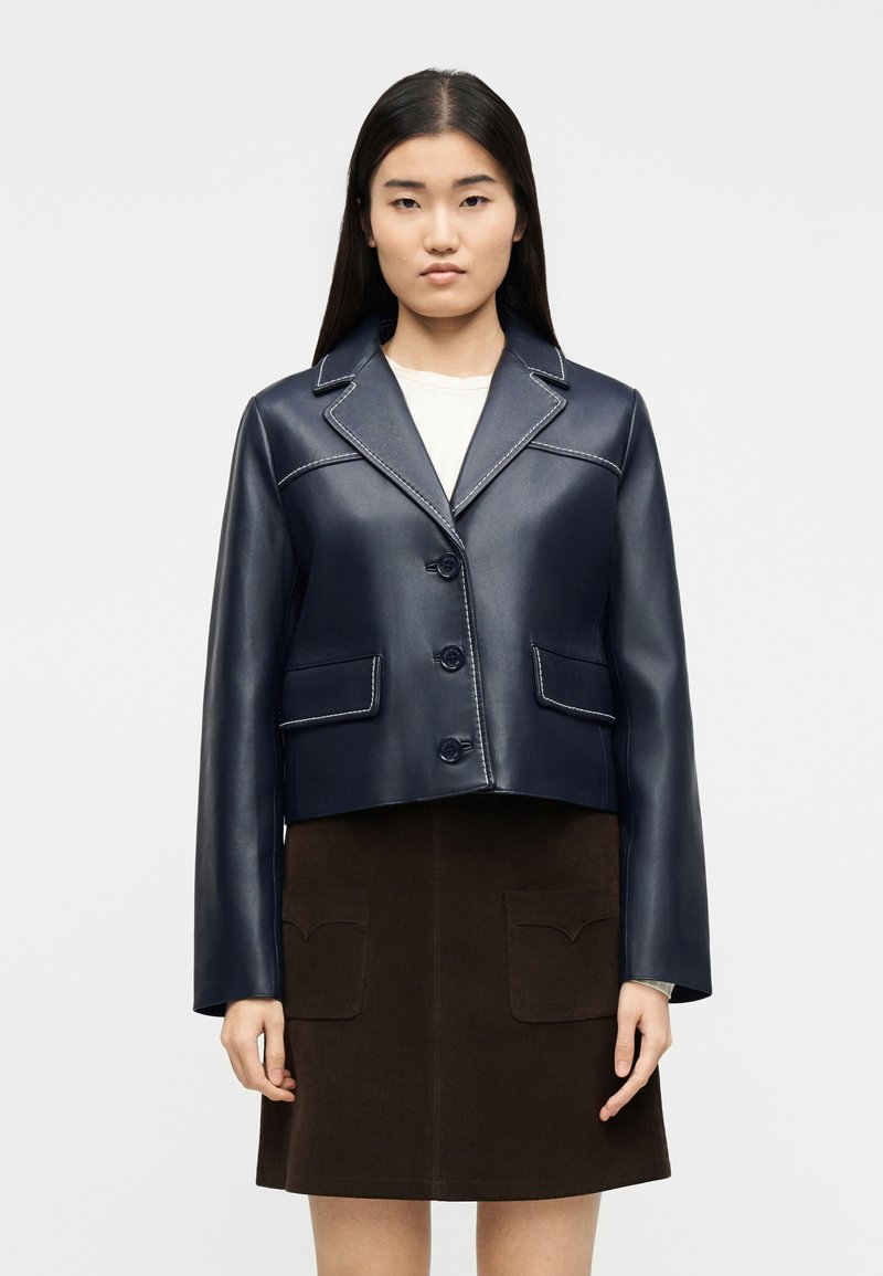 Femme portant une veste en cuir bleu marine boutonnée avec des coutures blanches sur un haut blanc et une jupe marron foncé avec des poches avant.