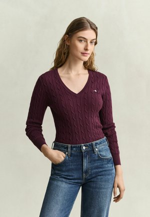 Vrouw met lang haar draagt een bordeauxrode kabeltrui met V-hals en blauwe spijkerbroek, staand met één hand in haar zak tegen een effen achtergrond.