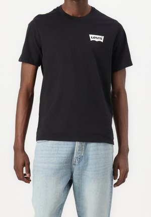 Homme portant un t-shirt Levi's noir uni et un jean bleu clair, debout devant un fond blanc.
