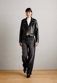 Zwart leren motorjack met zilveren hardware, witte coltrui, hoogsluitende zwarte jeans en zwarte schoenen op een houten vloer.