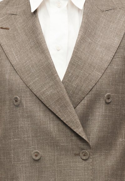 Μπεζ διπλής γραμμής σακάκι με υφή υφάσματος, τέσσερα κουμπιά καλυμμένα με ύφασμα, φορεμένο πάνω από λευκό πουκάμισο με γιακά και ορατά κουμπιά.