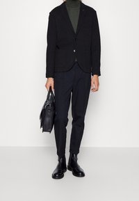 Svart, texturerad blazer med fickor, lager på lager över en mörkgrön polotröja. Kombinerad med mörka byxor och svarta, polerade ankellång stövlar. Läderväska ingår.