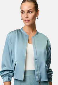 Helle blaue Satin-Bomberjacke mit gerippten Bündchen, Reißverschluss vorne und Seitentaschen. Über einem weißen Oberteil getragen, das eine glatte Textur aufweist.