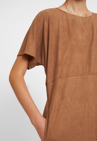 Braunes Wildleder-T-Shirt mit kurzen Ärmeln, rundem Ausschnitt und seitlichen Taschen. Glatte Textur, lässiges Design mit sichtbaren Nähten.