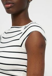 Κοντινό πλάνο ενός ατόμου που φοράει λευκό πουκάμισο με μαύρες οριζόντιες ρίγες, εμφανίζοντας τον ώμο, τον λαιμό και μερικό πρόσωπο σε απλό φόντο.