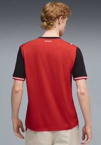 Αθλητική φανέλα κόκκινου και μαύρου χρώματος με κοντά μανίκια, με στρογγυλή λαιμόκοψη, οριζόντιες ριγέ λεπτομέρειες στα μανίκια και υφή στο ύφασμα.