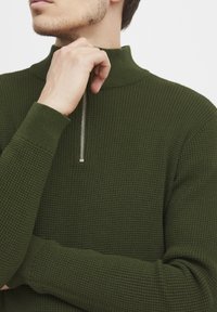 Pullover a maglia testurizzata di un verde scuro, con colletto alto e zip argentata. I polsini aderenti e la vestibilità rilassata mettono in risalto il motivo del materiale.