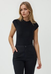 Schwarzes, enges T-Shirt mit kurzen, angeschnittenen Ärmeln und rundem Ausschnitt. Getragen mit schwarzen Hosen, die aus einem glatten, dehnbaren Stoff bestehen.