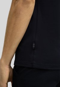 Czarna koszulka sportowa z teksturowanej tkaniny, z widocznym szwem bocznym i małą metką z logo blisko dołu. Materiał wydaje się oddychający i lekki.