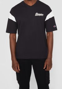 Camiseta negra de manga corta con escote en pico y acentos blancos en los hombros, con un logotipo de "Champion" en el pecho y un logotipo cosido en la manga.