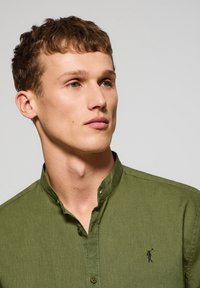 Olijfgroene, langmouwen shirt met een mandarijnkraag, knoopsluiting en een klein zwart geborduurd logo op de borst. Glad stoftextuur.