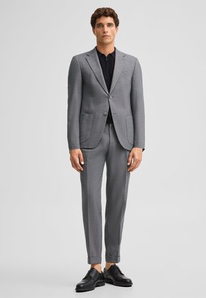 Uomo che indossa un completo grigio con camicia nera e scarpe eleganti nere, in piedi davanti a uno sfondo grigio chiaro uniforme.