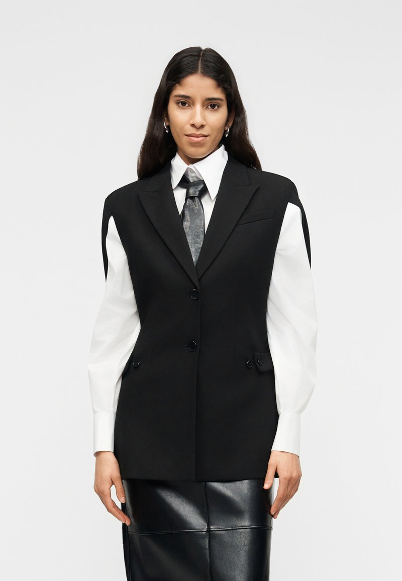 Γυναίκα με μακριά σκούρα μαλλιά που φοράει υπερμεγέθη μαύρο σακάκι, λευκό πουκάμισο με φουσκωτά μανίκια, ασημένια γραβάτα και μαύρη δερμάτινη φούστα.