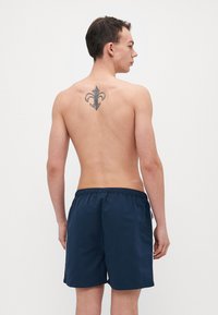 Marineblå badebukser med elastisk liv, glat tekstur og en sideslids. En sort fleur-de-lis tatovering er synlig på ryggen.