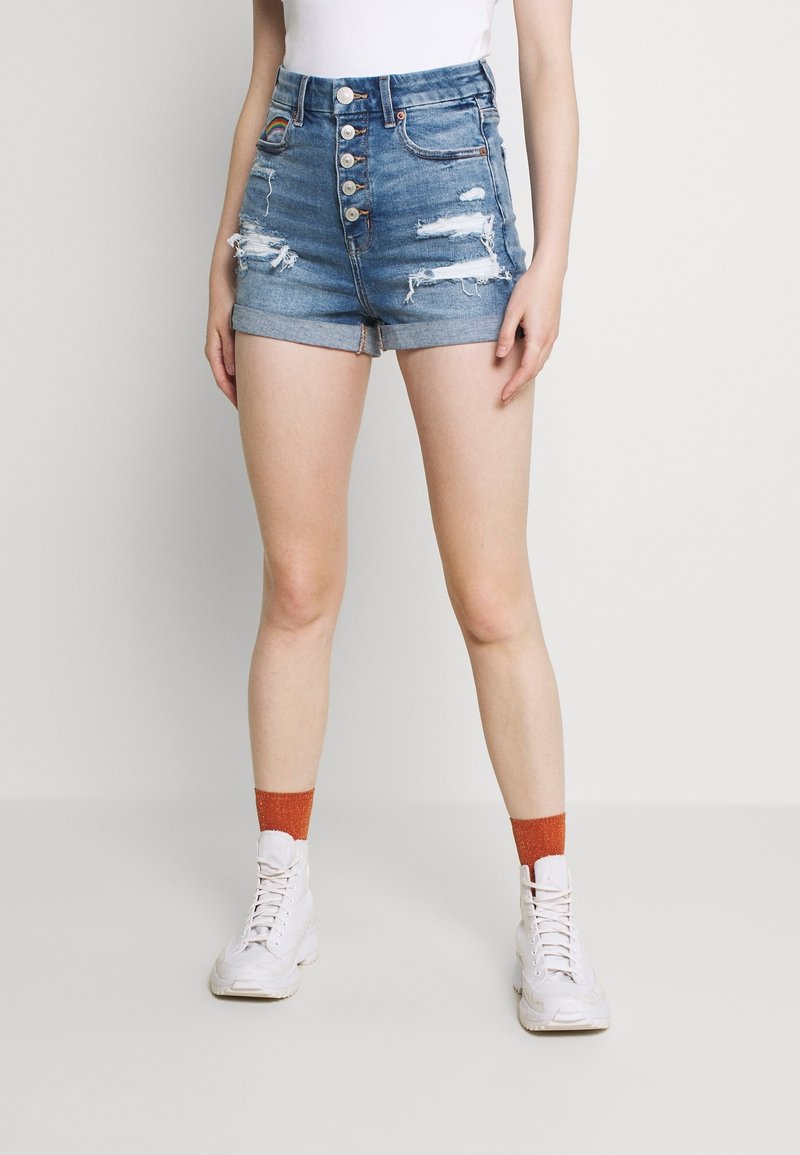 Femme portant un short en jean déchiré taille haute avec une braguette à boutons, des chaussettes orange, des baskets blanches et un haut blanc rentré, sur un fond uni.